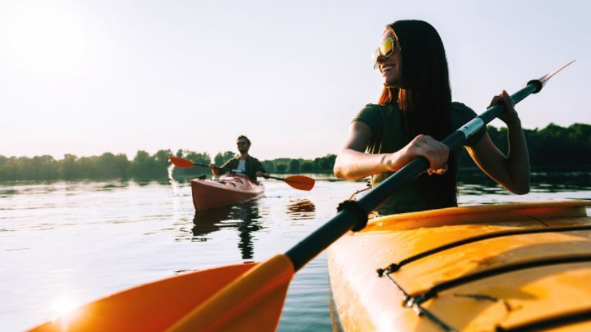 Woman kayaking on a lake, holding paddle. Another kayaker in background. Sunny, orange glow.