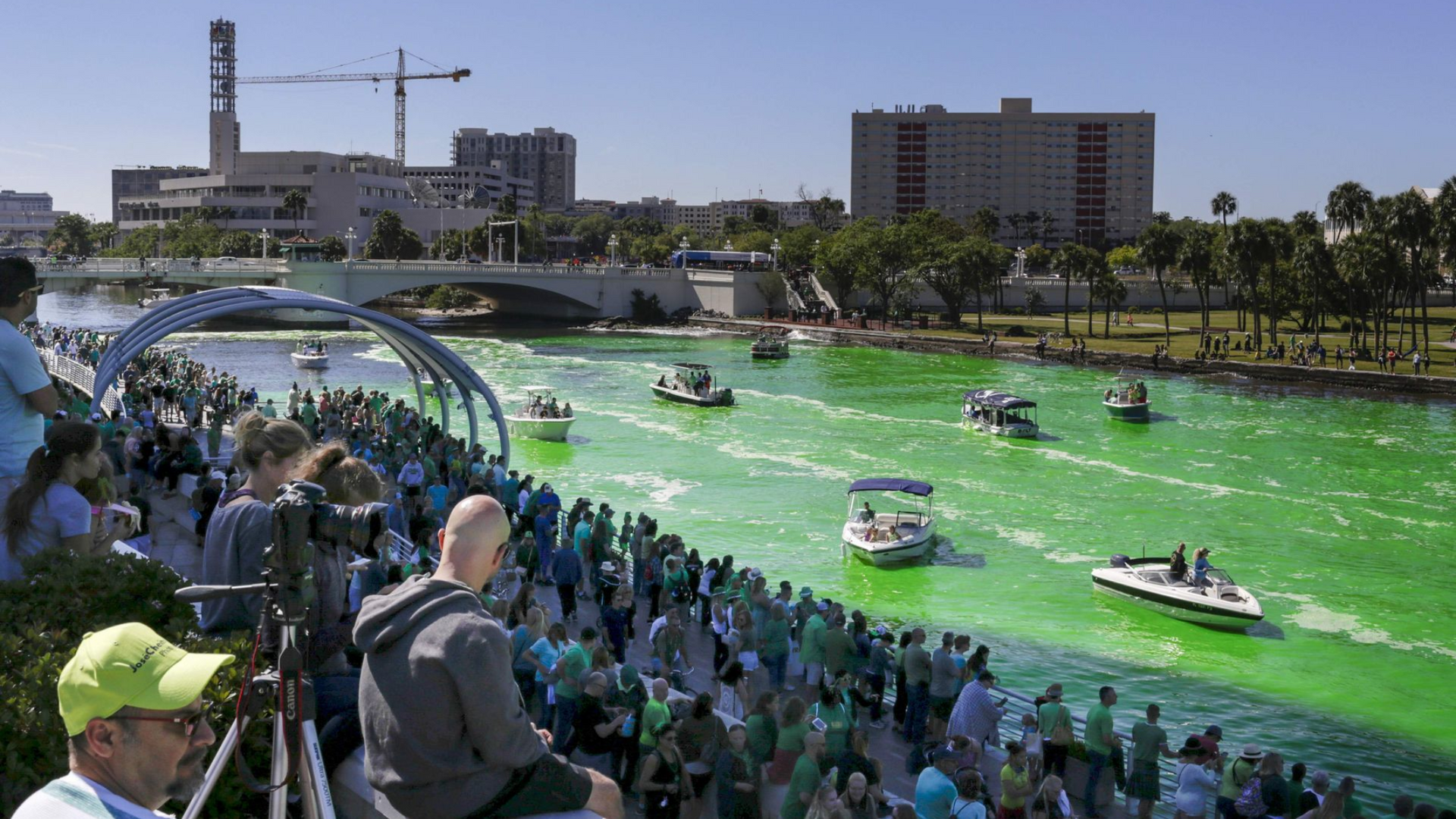 Green river with boats, surrounded by crowds of people along the waterway. St. Patrick's Day celebration.