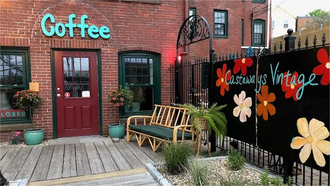 Goffee shop entrance with red door, teal sign, and vintage decor, including a bench and flower-decorated fence.