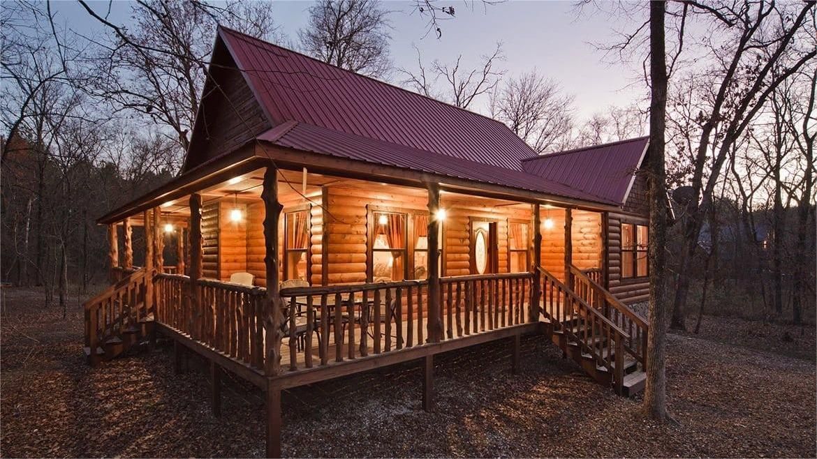 Wooden cabin with a red roof and porch, lit from within, surrounded by trees.