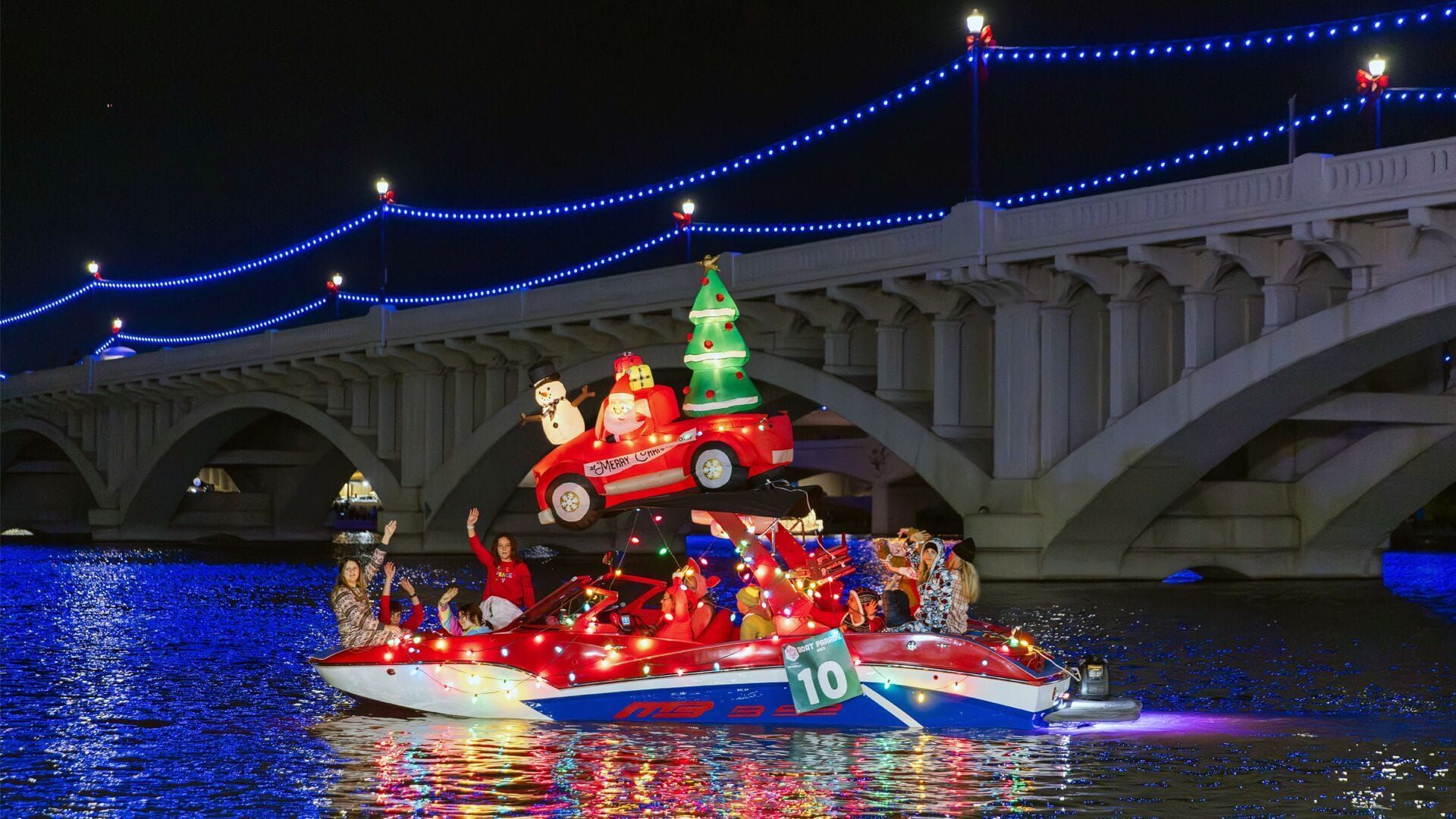 A boat decorated with Christmas lights and Santa waving, with a bridge in the background.