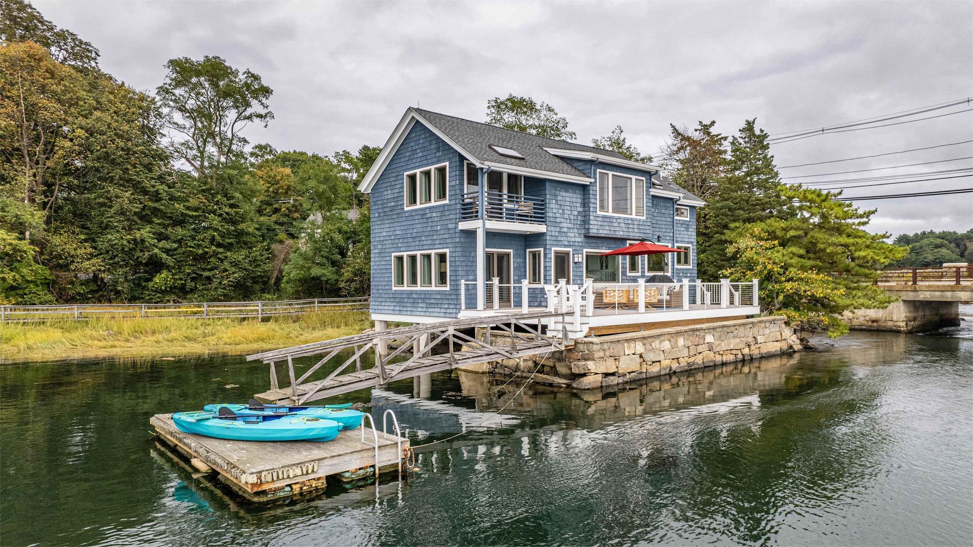 Blue house on a floating dock with a kayak, overlooking a waterway with a bridge.