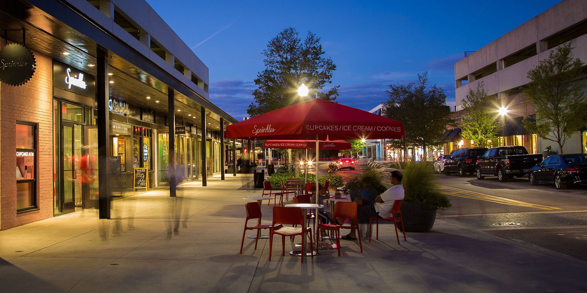 Nighttime outdoor dining area with red umbrellas, tables, and chairs. Buildings and parked cars in the background.