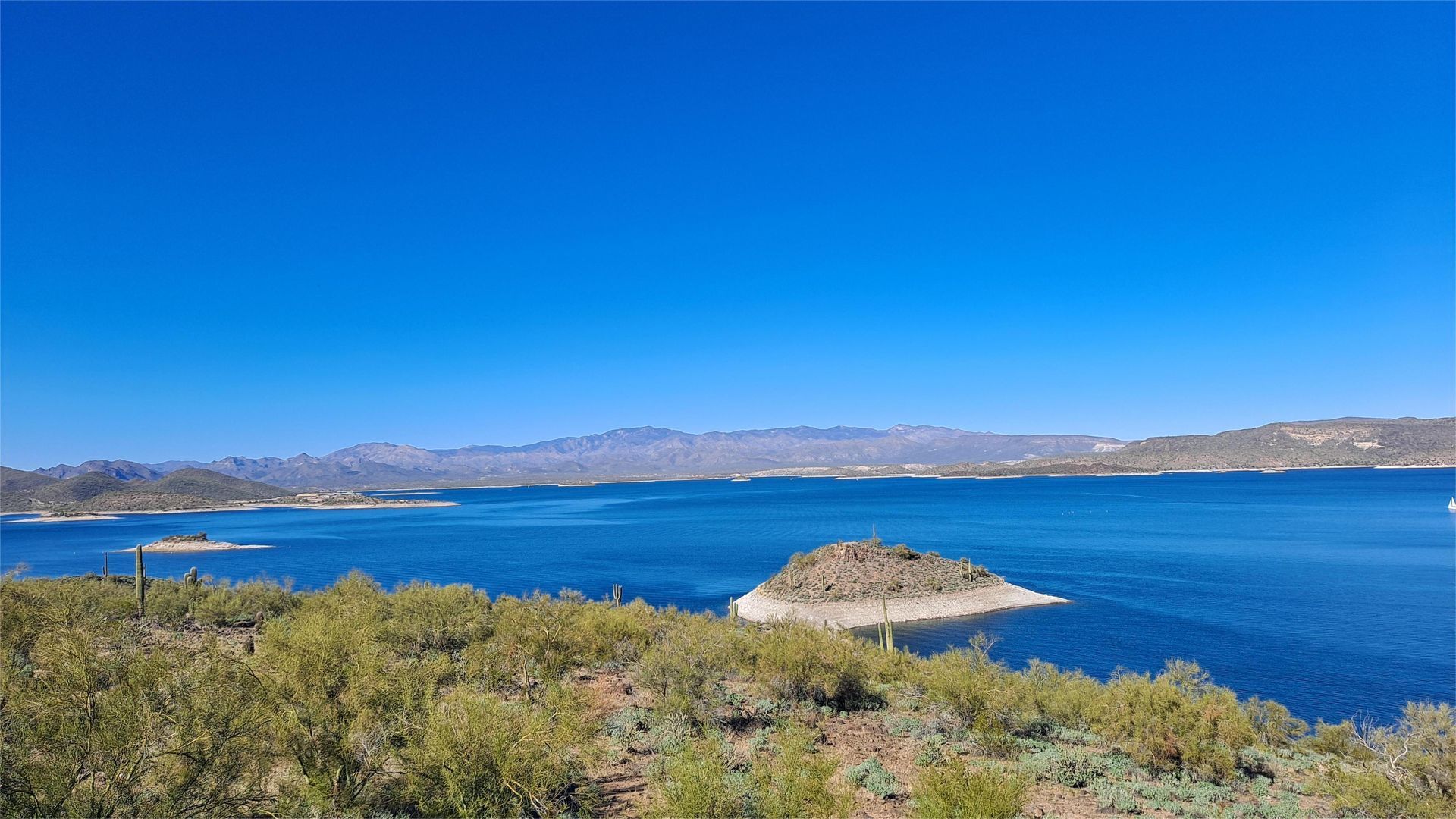 Blue lake with small islands and distant mountains under a clear blue sky.