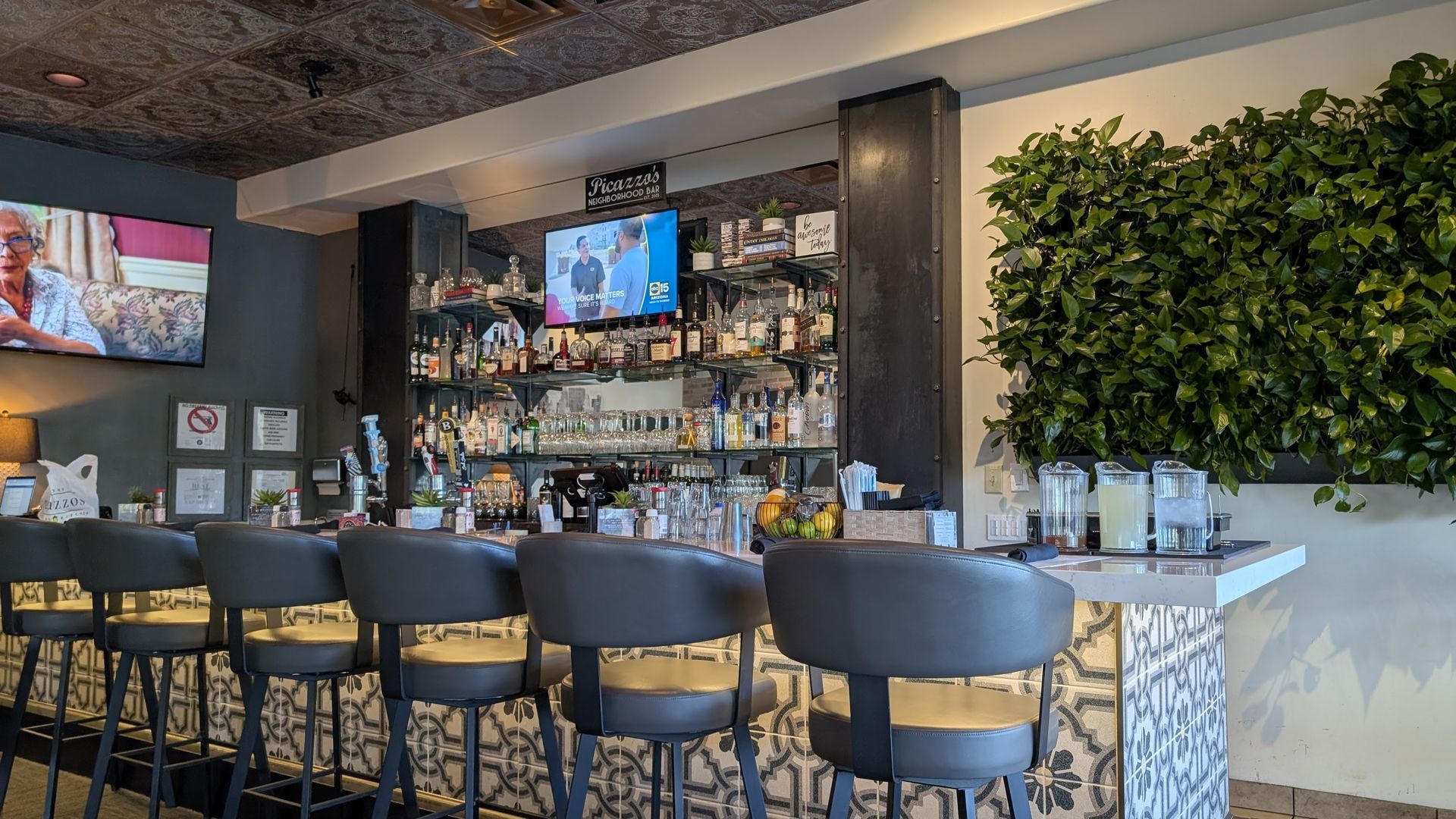 Bar interior with barstools, liquor bottles, a TV, and a wall of greenery.