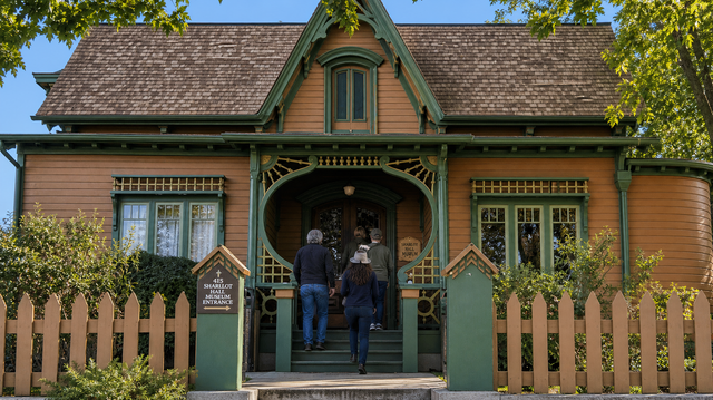 Victorian-style house with green trim, porch, picket fence, and two people standing at the front door