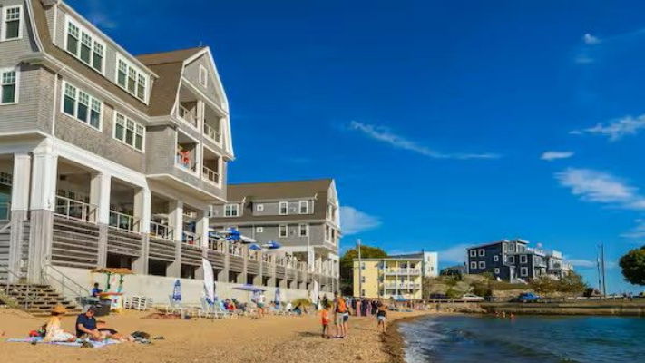 Beach scene with gray buildings, blue sky, and people on the sand near the water.