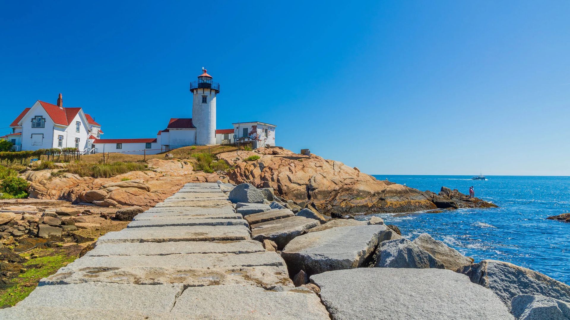 Lighthouse on rocky coast under a blue sky; broken stone path in foreground leads to the ocean.