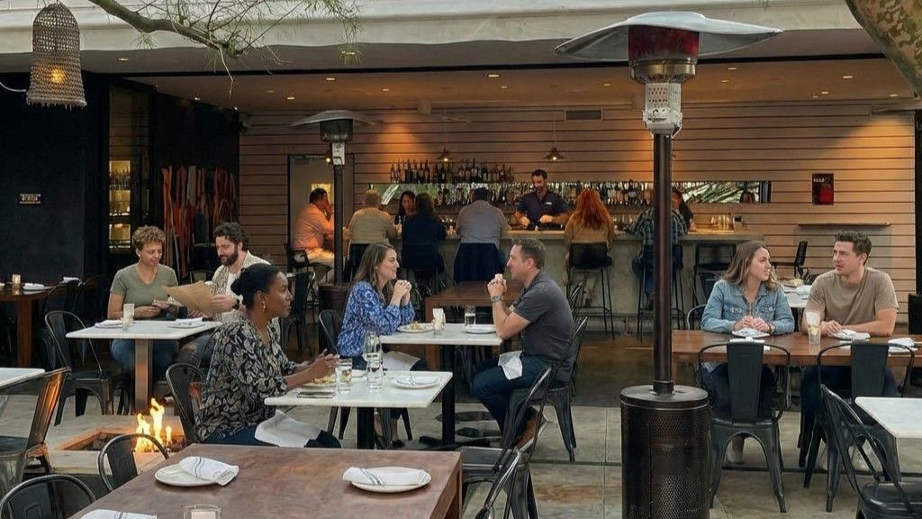 Patrons dine at tables in a modern, dimly lit outdoor restaurant patio featuring a fireplace and space heaters.