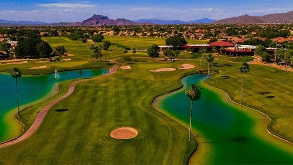 Aerial view of a green golf course with blue water features, palm trees, and a clubhouse against a desert mountain backdrop.
