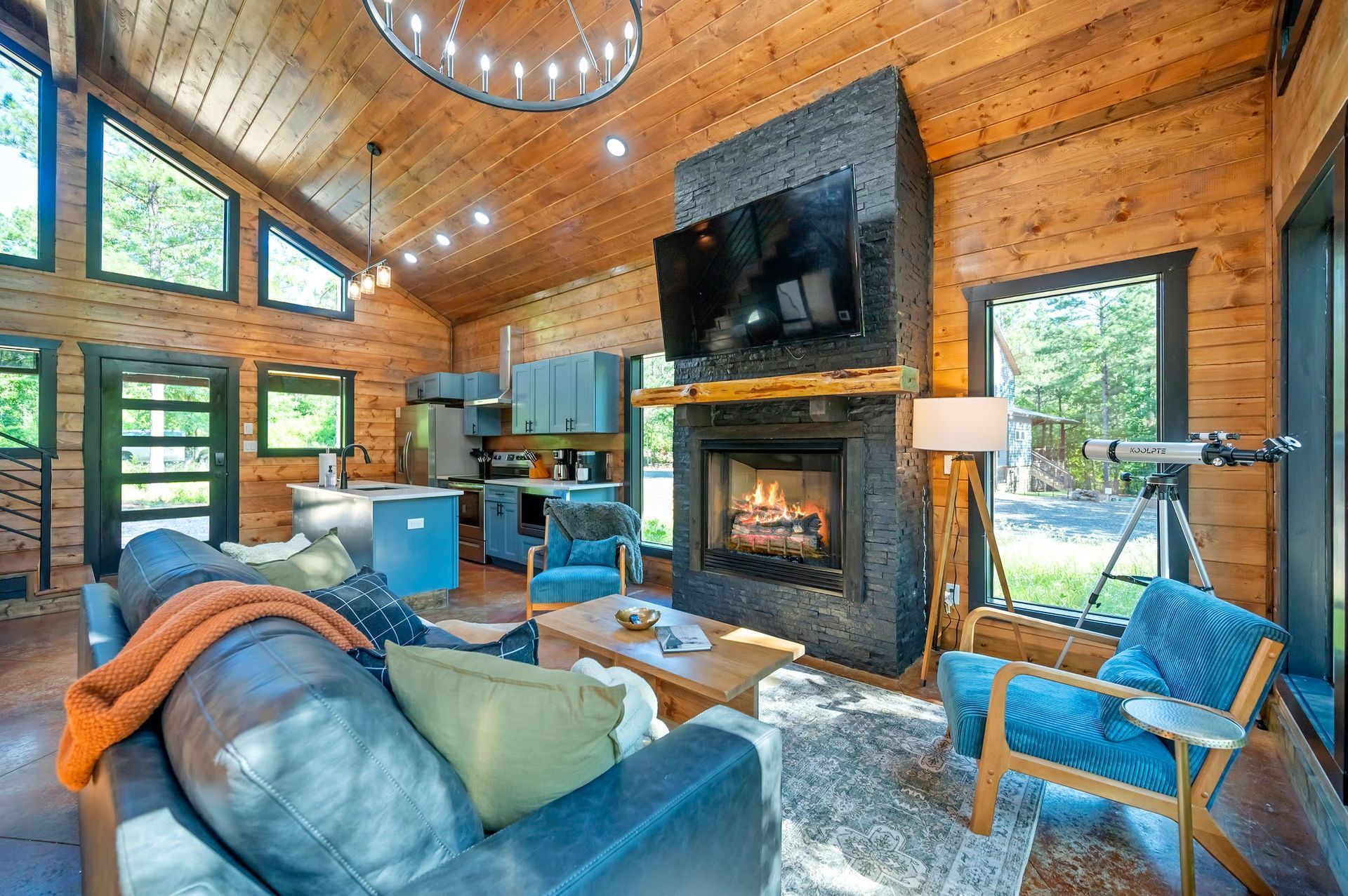 Rustic living room with stone fireplace, wood-paneled walls, large windows, and blue chairs around a sofa.