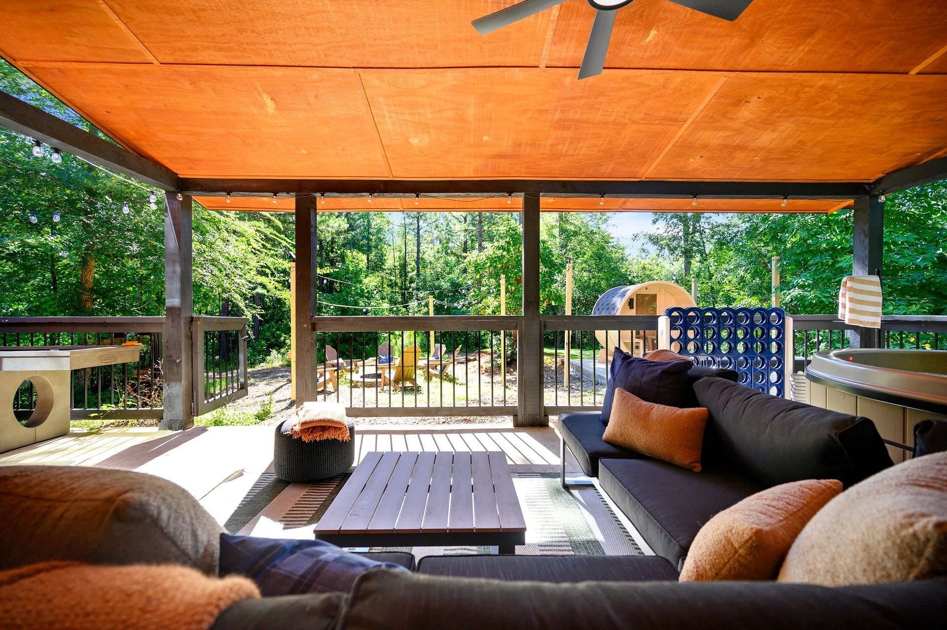 Covered outdoor deck with cushioned seating, wooden coffee table, and lush green trees beyond the railing