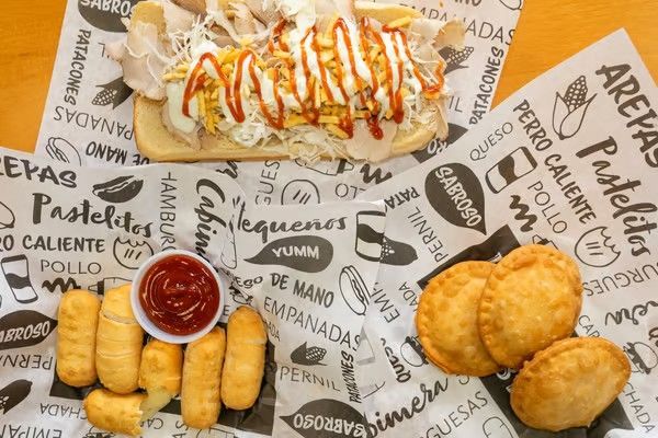 A table with a sandwich, empanadas, tequeños, and ketchup on patterned paper with food terms.