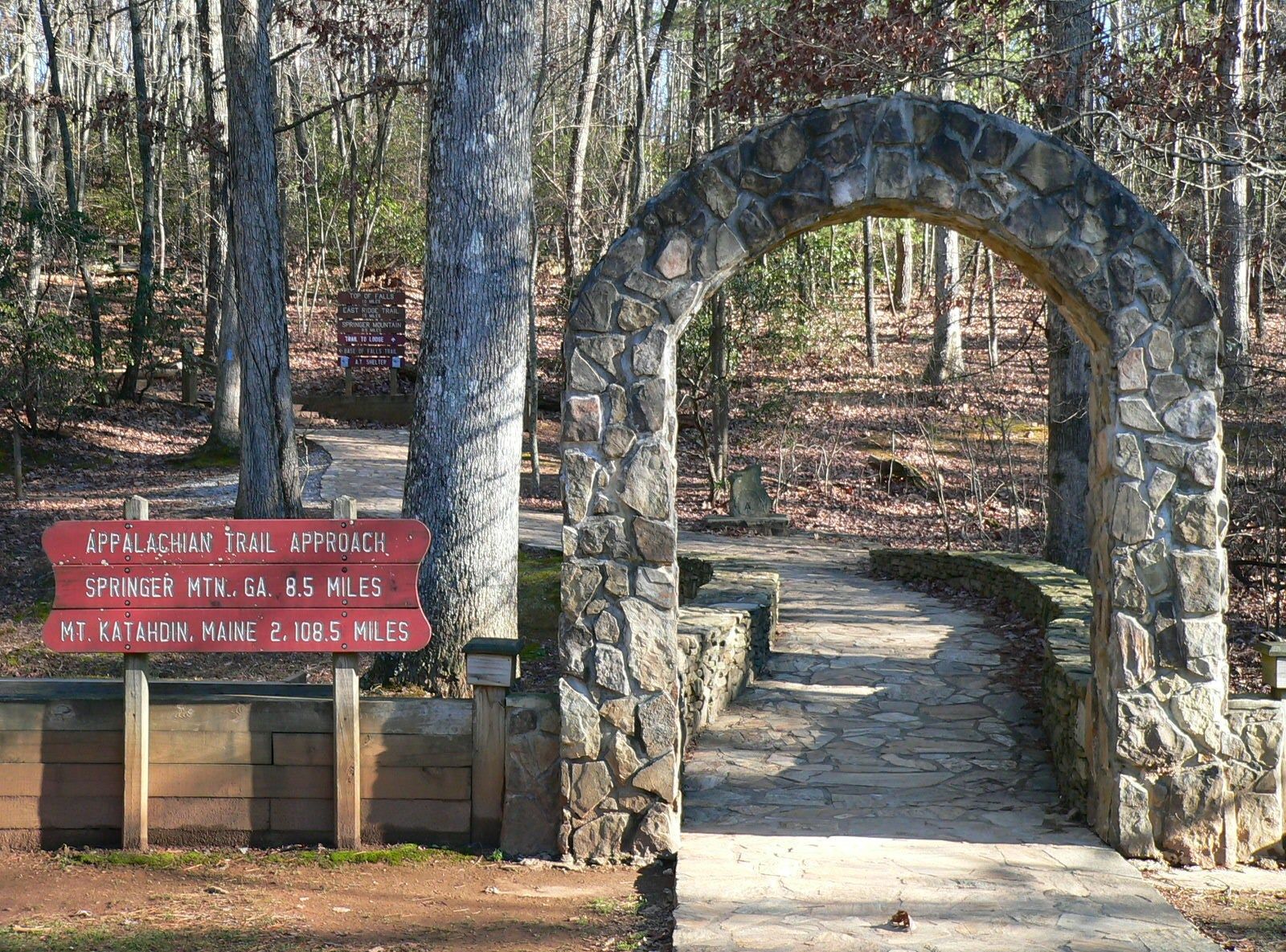 Stone archway over a path in a wooded area. A sign on the left has red lettering.