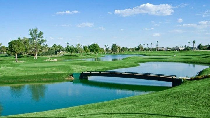 Green golf course with a bridge over water under a blue sky with clouds.