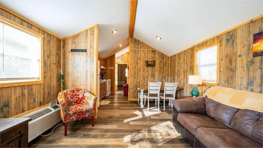 Cozy cabin living room: wood-paneled walls, sofa, patterned armchair, dining table, windows.