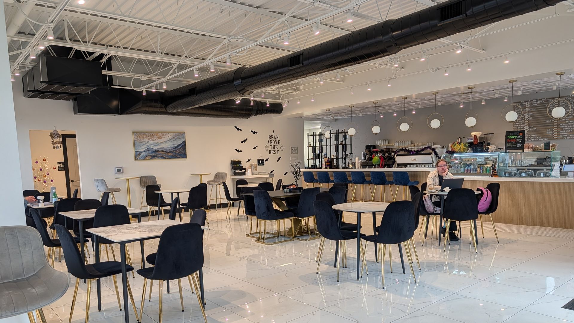 An interior view of a bright cafe with black chairs, tables, and an open bar area against a long counter.