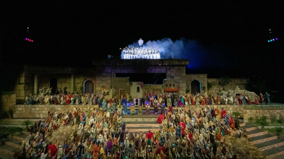 Large crowd of people in colorful costumes, gathered on tiered steps with a stage set at night.