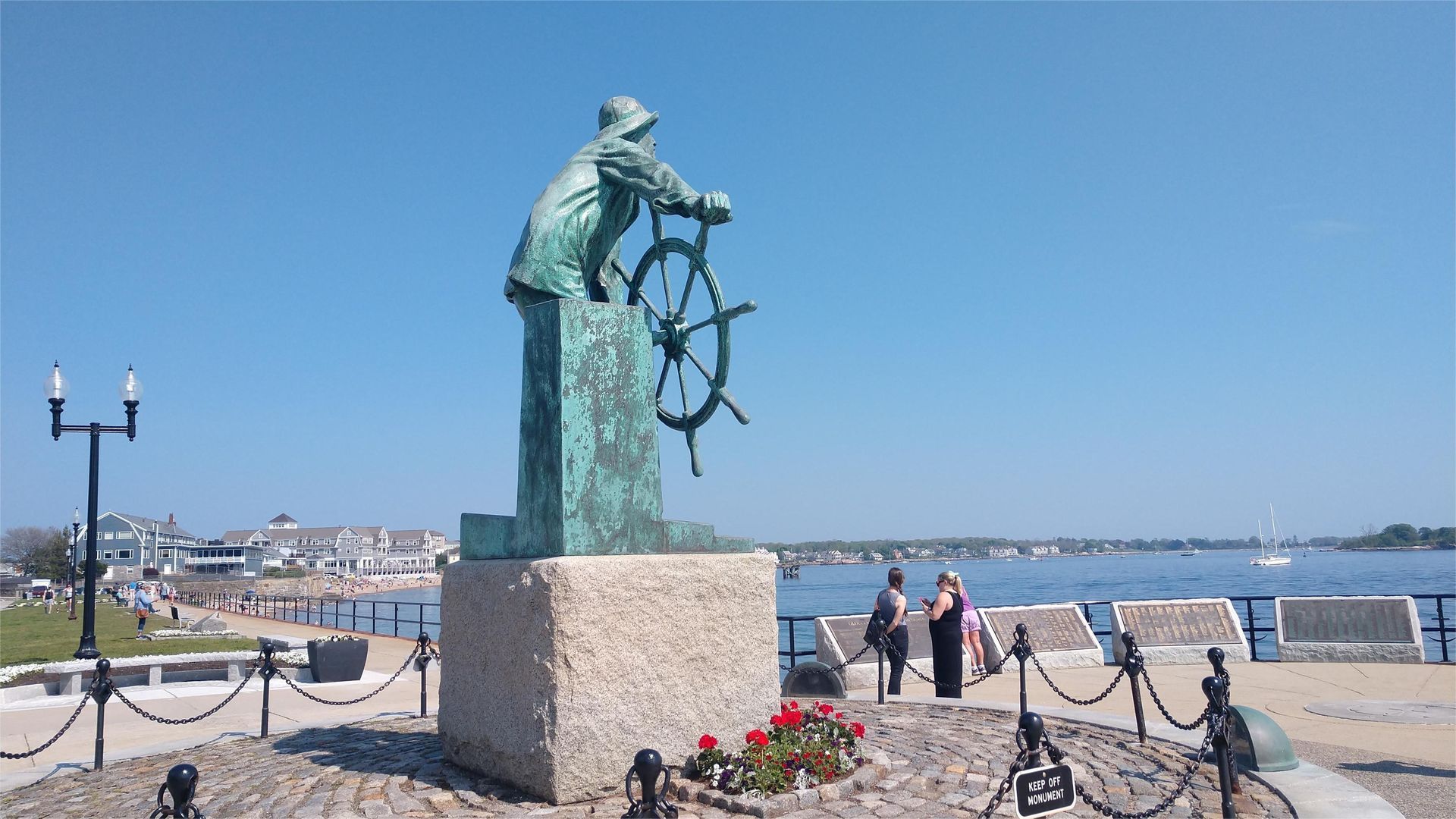 Statue of a sailor at the helm, overlooking the water under a blue sky.