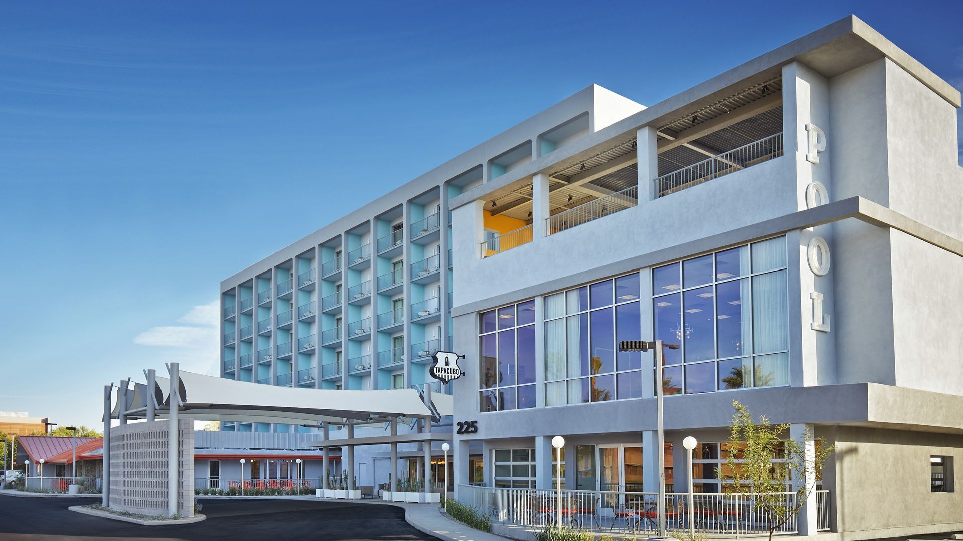 A modern, white hotel building with large windows and a covered entrance under a clear, blue sky.