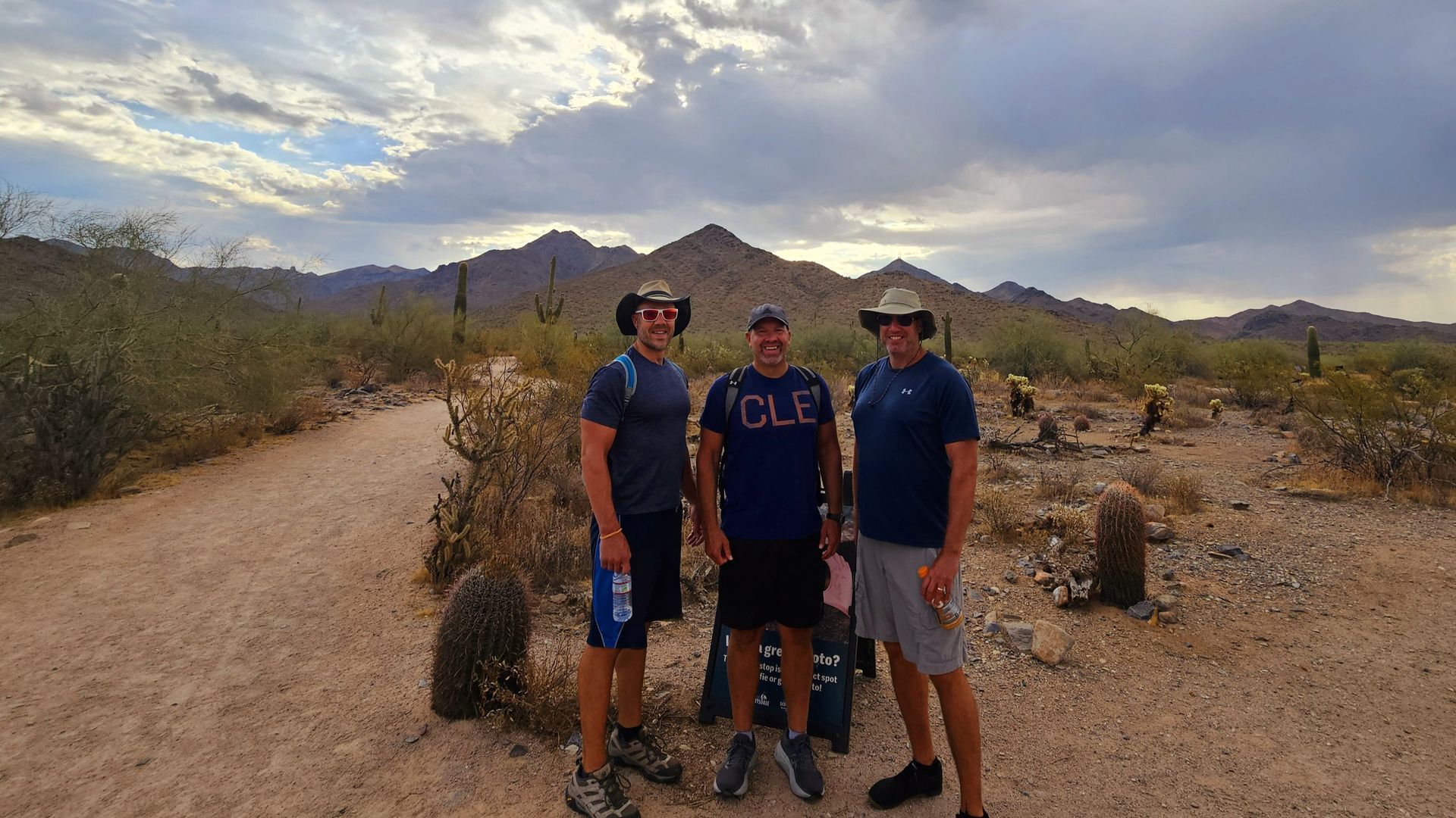 Three people stand on a desert trail with mountains in the background under a cloudy sky.