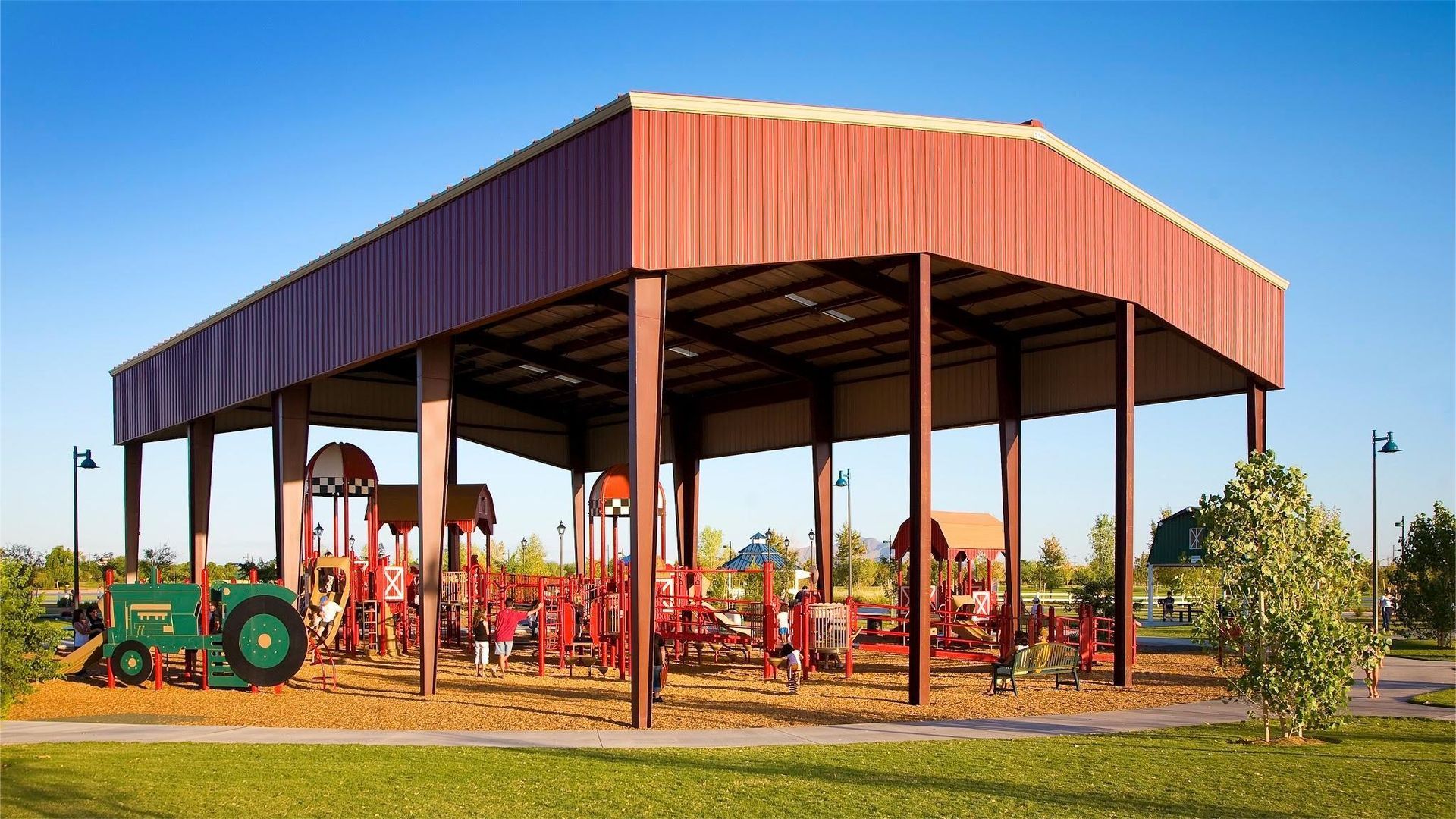 Red shelter over playground with farm equipment. Green tractor, brown poles, and blue sky.