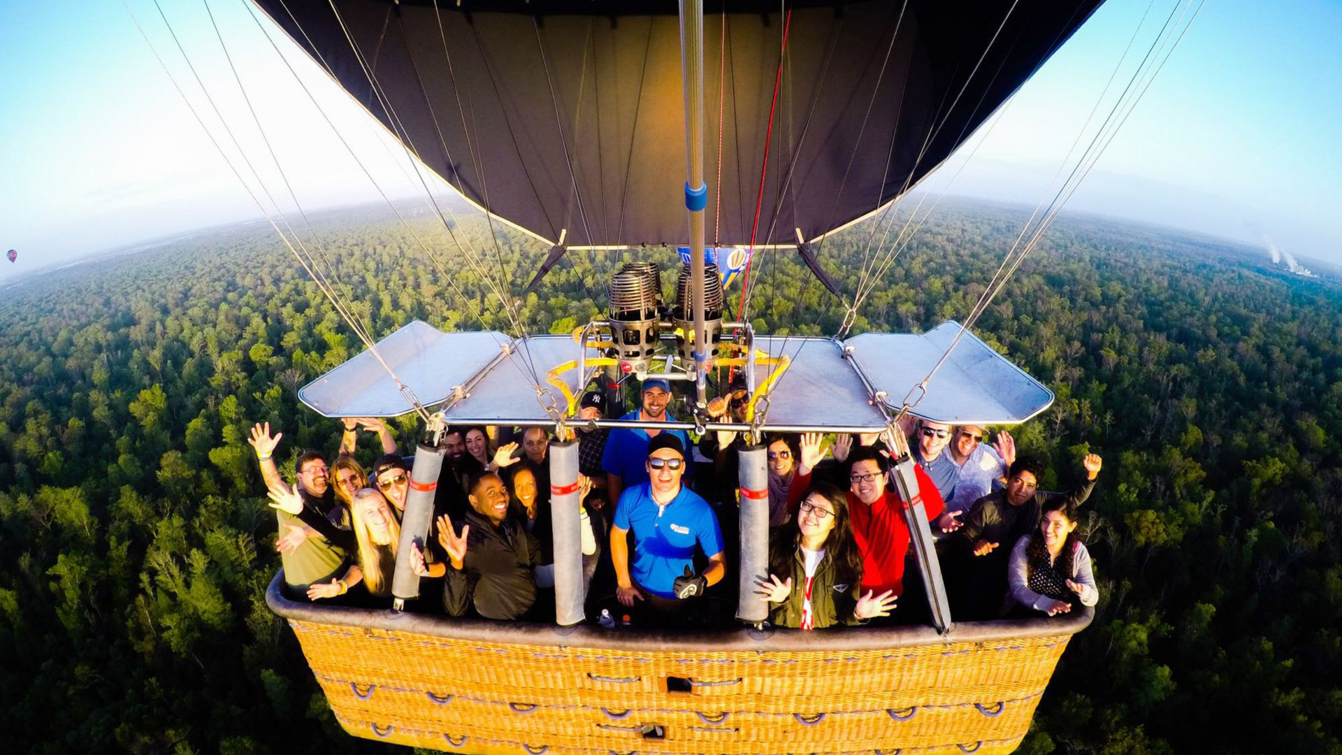 People in a hot air balloon basket flying over a forest, many are waving.