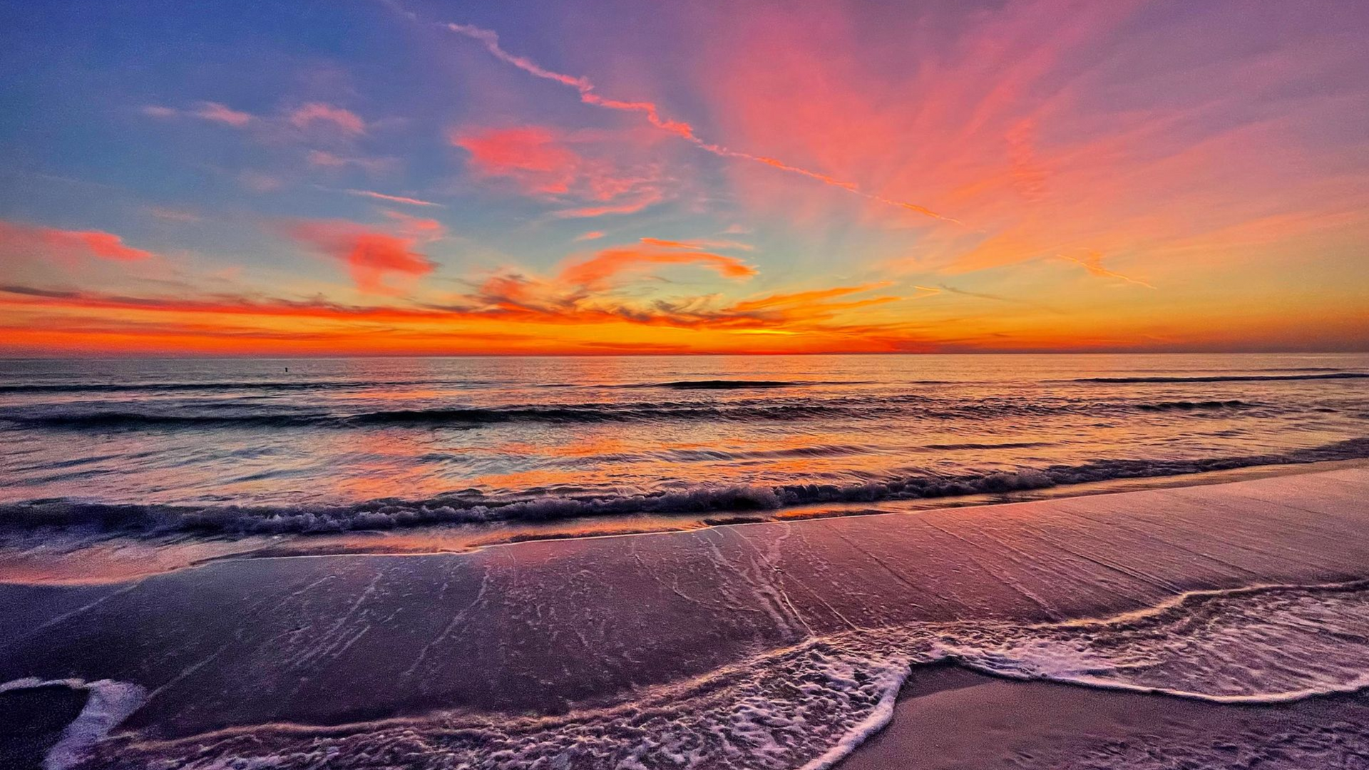 Vibrant sunset over ocean; orange and pink sky reflects on water. Waves gently crash on beach.