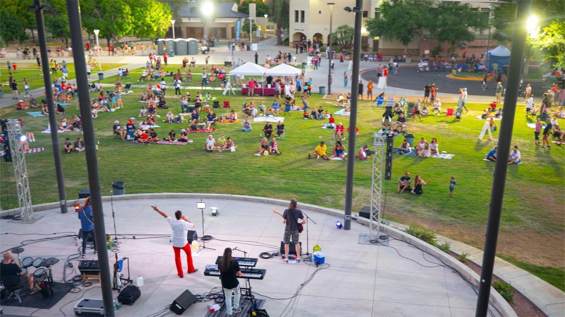 Band performing on a stage with a crowd on a grassy lawn. Dusk, lights, and buildings in background.