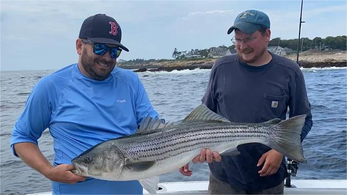 Two men on a boat hold a large striped bass fish. Blue water and coastline in the background.