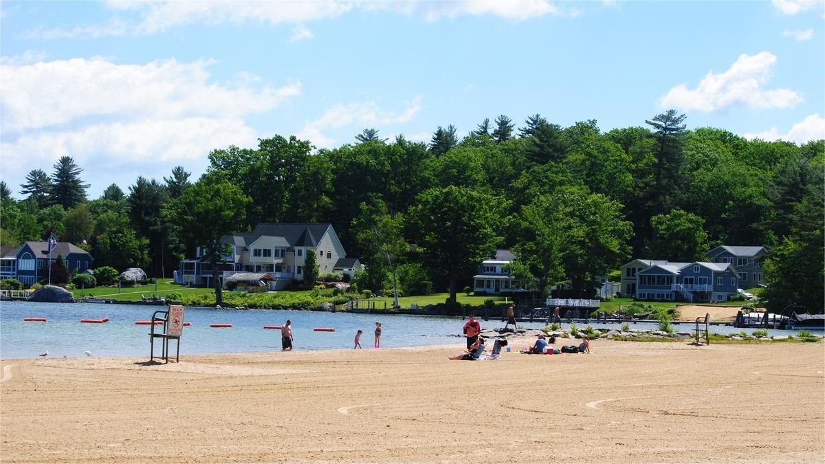 Sandy beach with people near water, houses and trees on a sunny day.