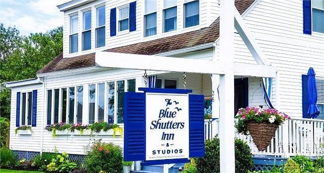 Blue Shutters Inn & Studios sign in front of a white house with blue shutters and a hanging flower basket.