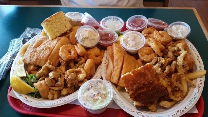 Two loaded seafood platters on a red tray: fried fish, shrimp, clams, fries, sauces, and cornbread.