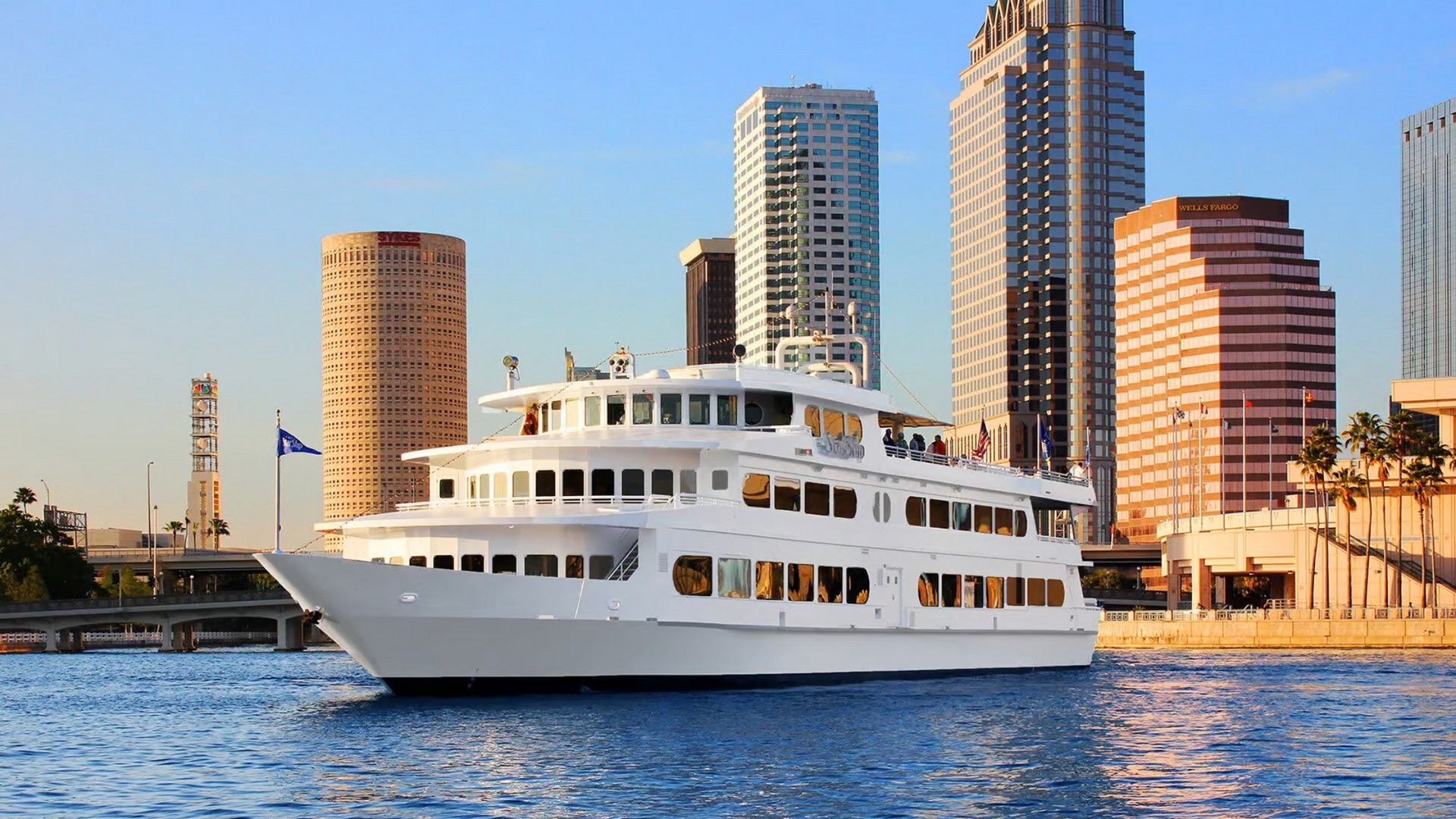 White multi-level boat on water, city skyline in background, blue sky.