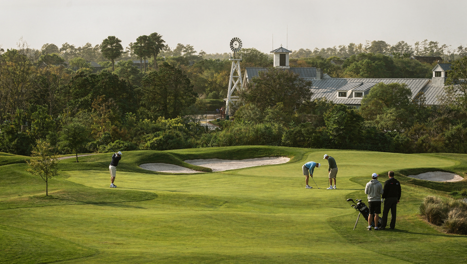 Golfers on a green course near sand bunkers and white houses in the background.