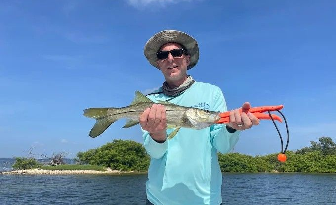 Man holding a snook fish on a boat; blue sky, water, and green island in the background.