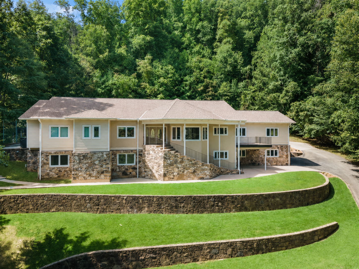 Large beige house with stone foundation on a grassy hillside, surrounded by dense green trees