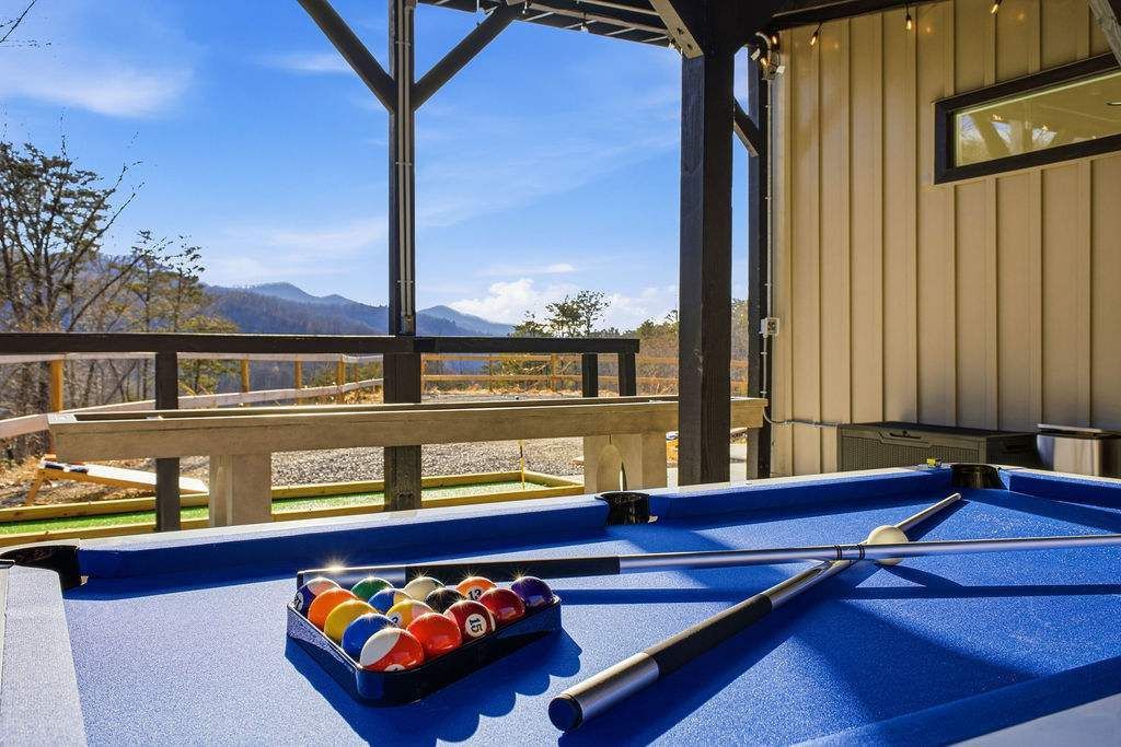 Blue pool table under a covered patio, with balls racked and cue sticks beside a scenic outdoor view