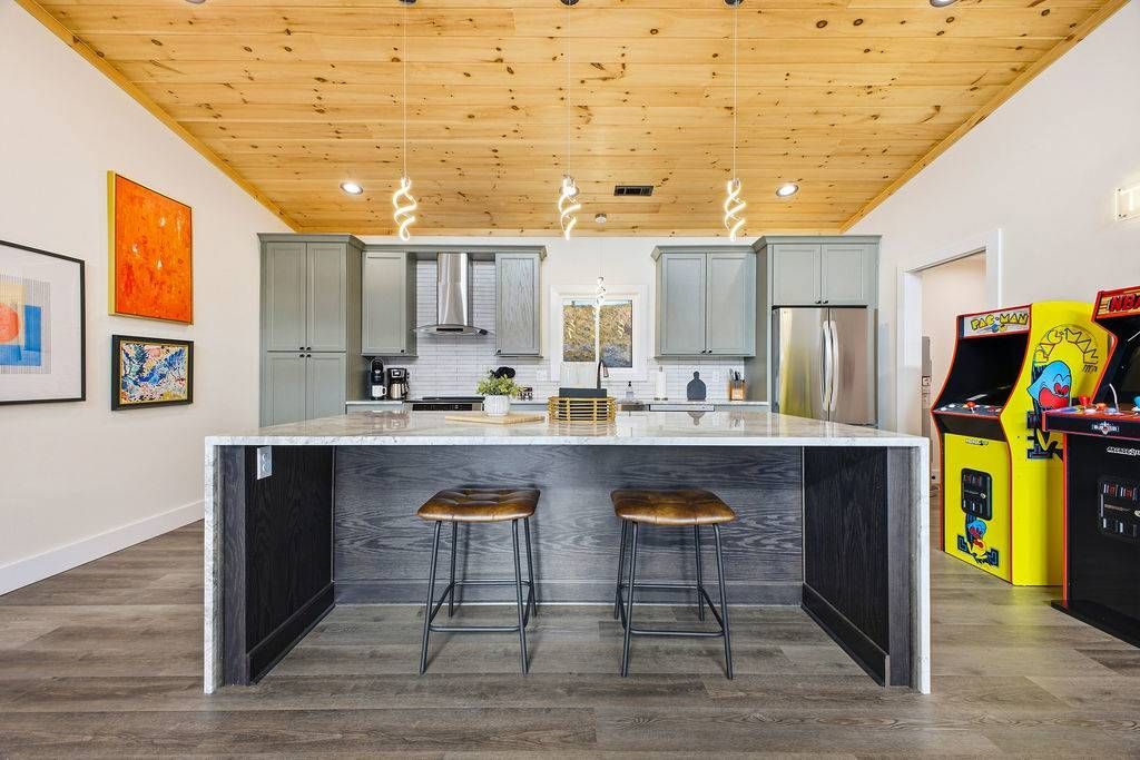 Modern kitchen with island, two stools, wood ceiling, stainless appliances, and colorful arcade games on the right