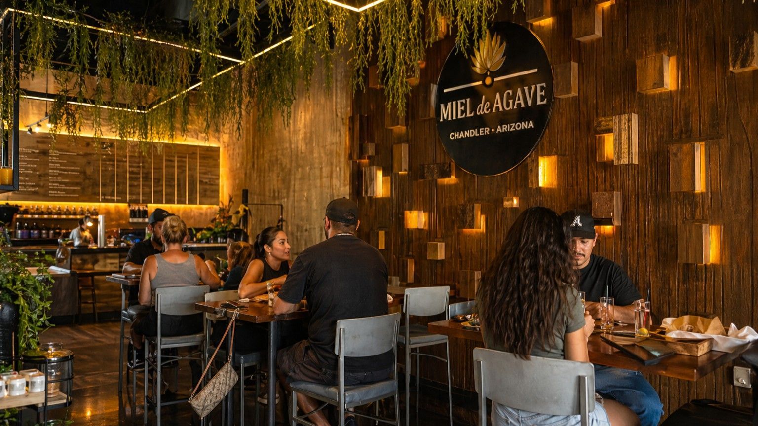 Cozy restaurant interior with warm amber lighting, hanging greenery, and diners seated at wooden tables.