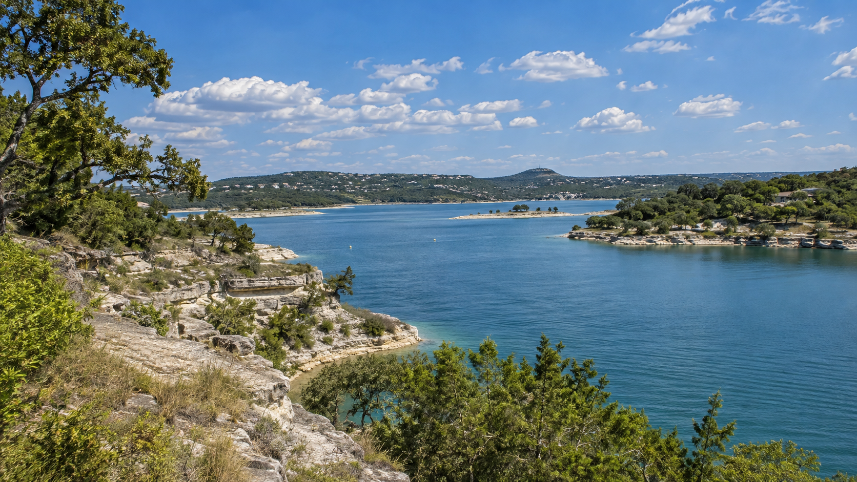 Rocky lakeshore with blue water, green trees, and distant hills under a partly cloudy sky