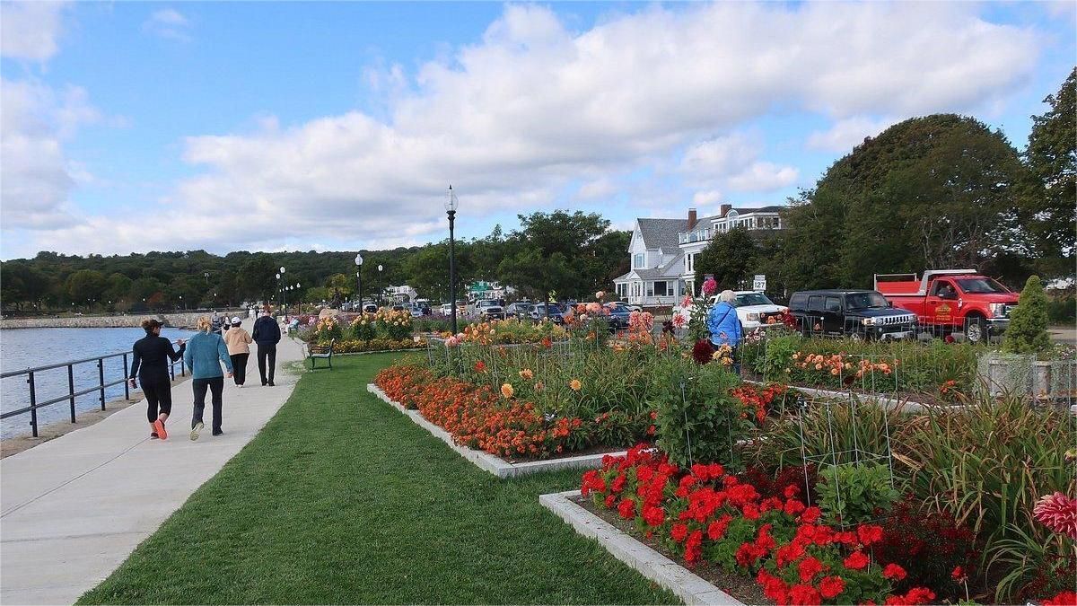 People walking on a waterfront path beside flower beds and parked cars under a cloudy sky.