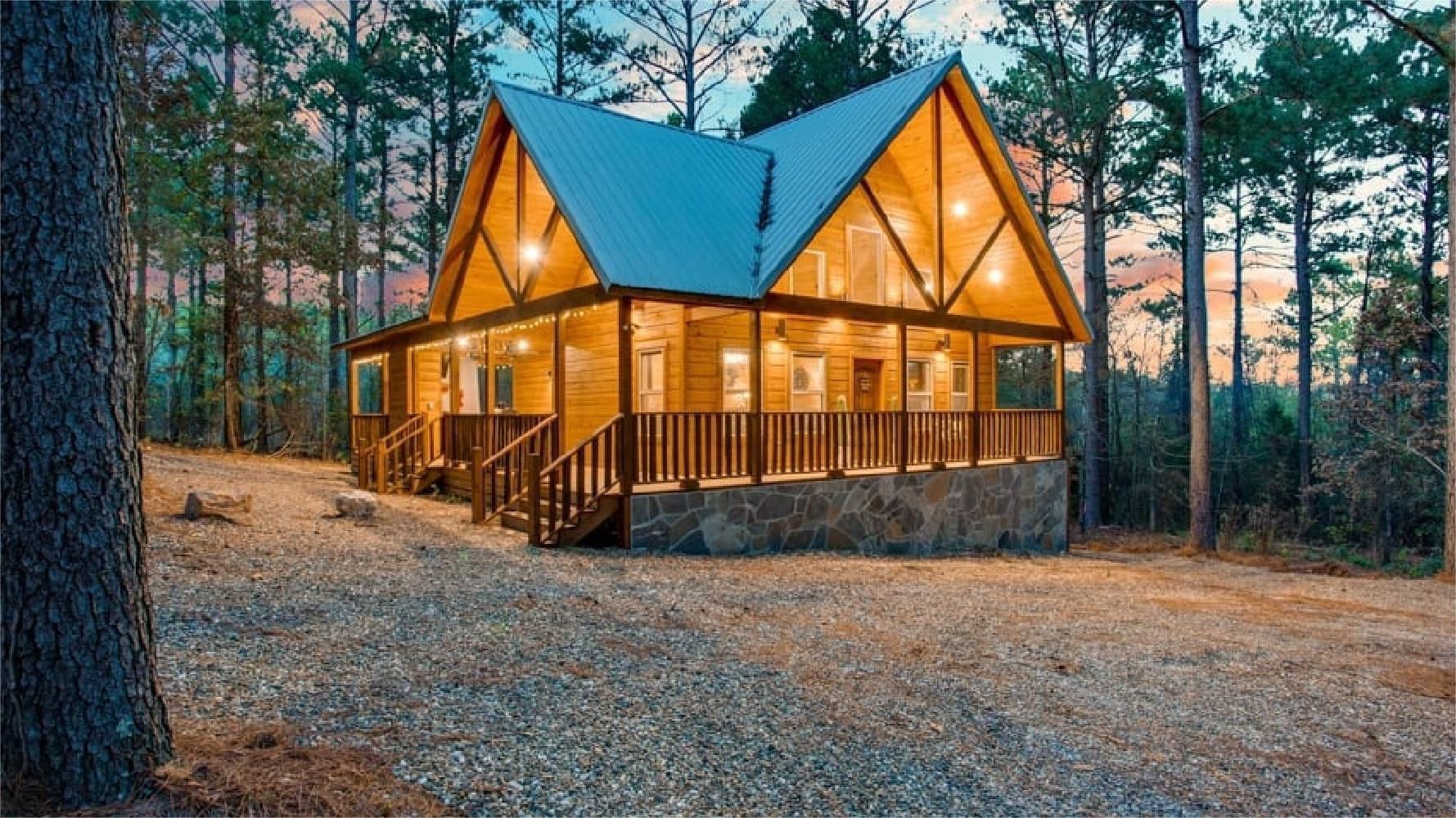 Wooden cabin with large windows and porch, nestled in a forest at dusk.