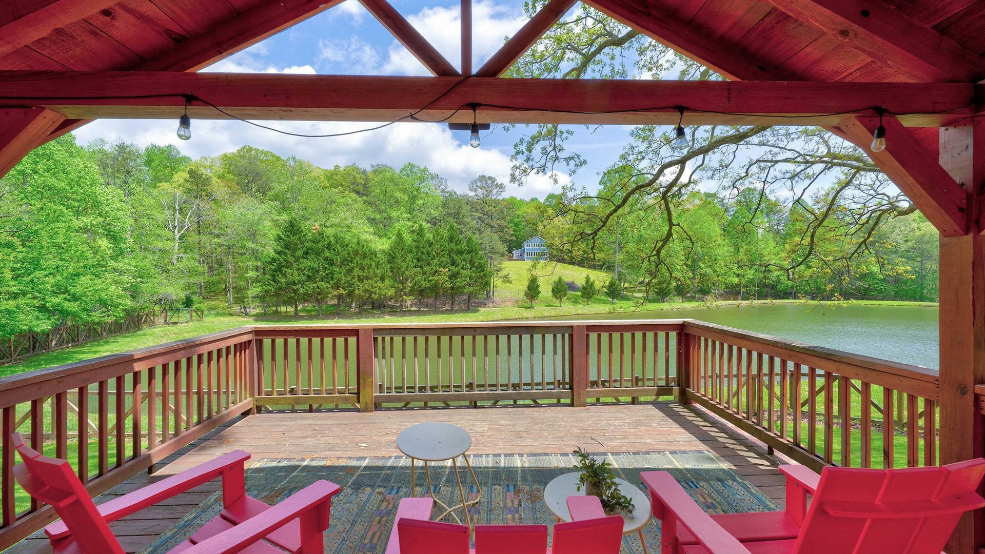 Red patio chairs sit on a deck under a wooden pavilion overlooking a lake surrounded by green trees.