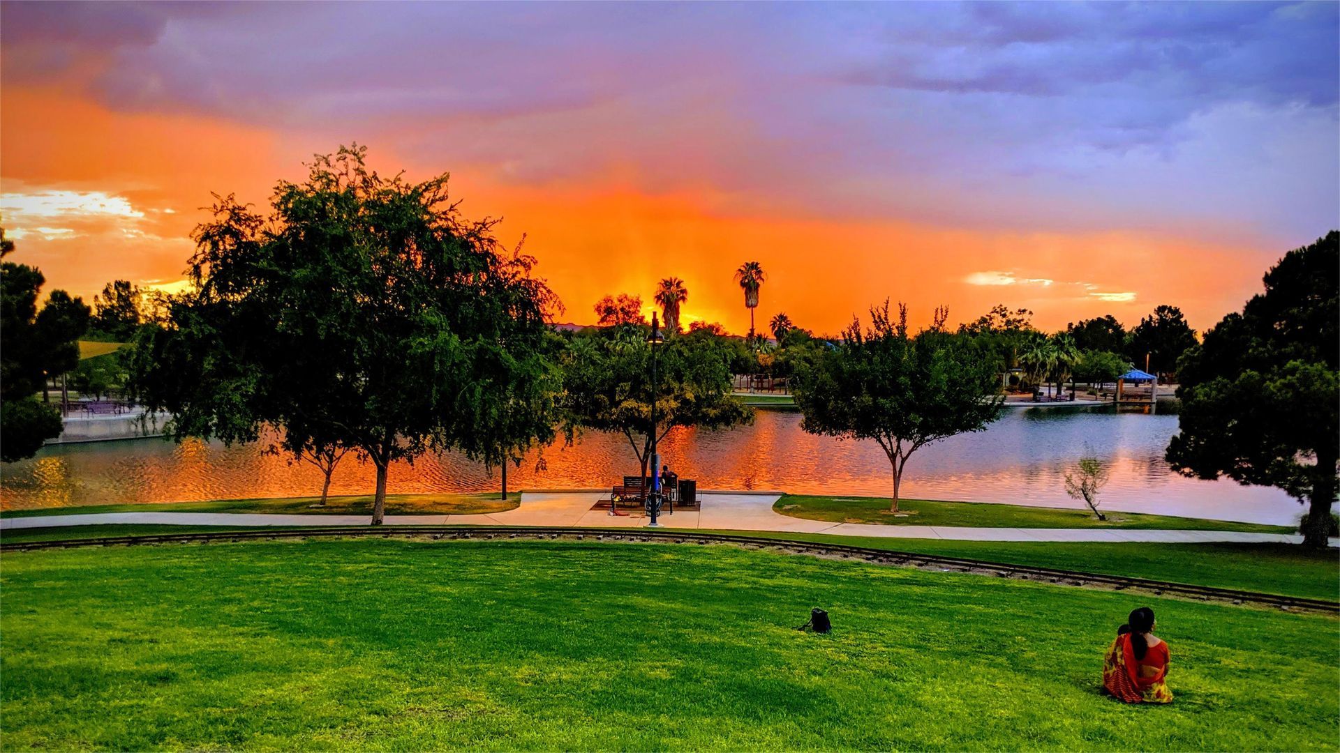 Vibrant sunset over a park lake, green grass in foreground, trees silhouetted, orange and purple sky.