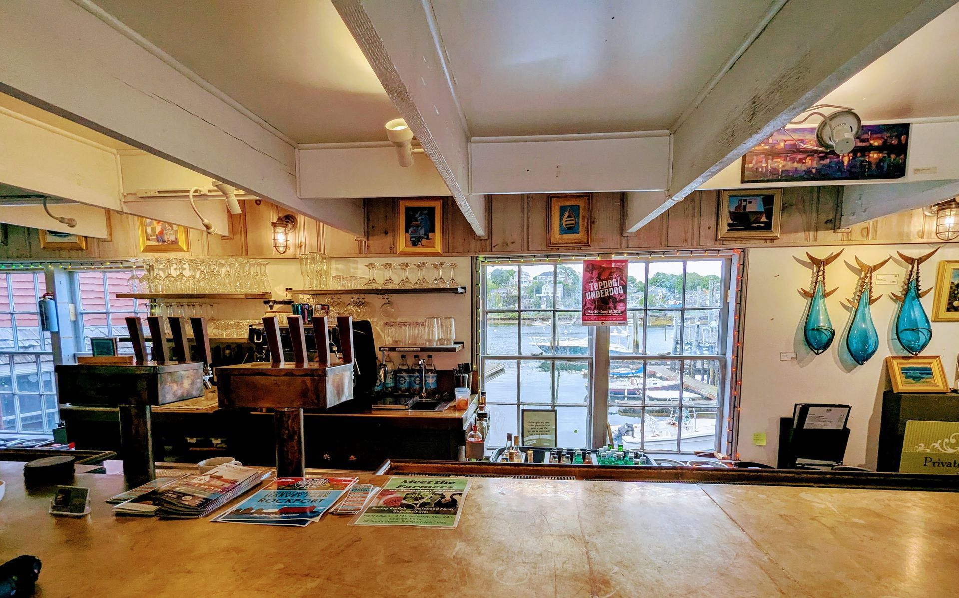 Interior of a restaurant with a bar, windows overlooking water, and fishing-related artwork.