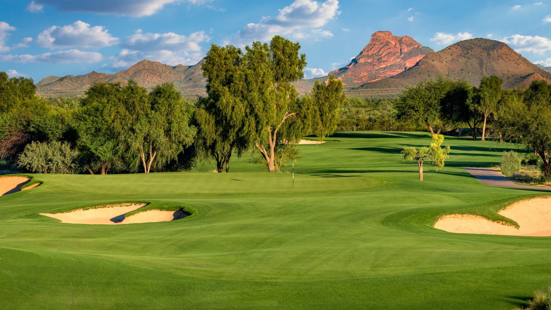 Green golf course with sand traps and trees, mountains in the background under a blue sky.