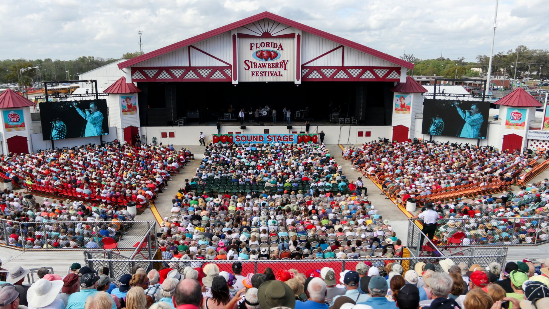 Large outdoor concert stage with a packed audience. Red and white building. People seated, watching.