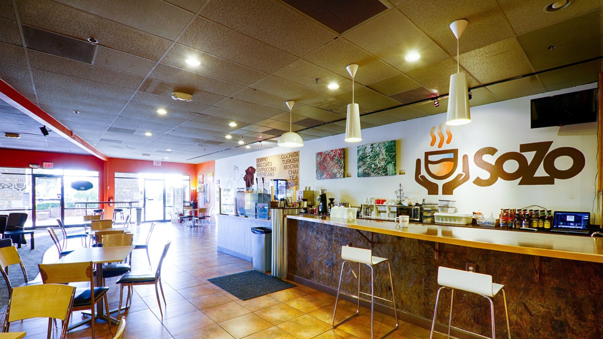 Interior of Sozo cafe: Bar with stools, tables, chairs, and logo on a white wall.