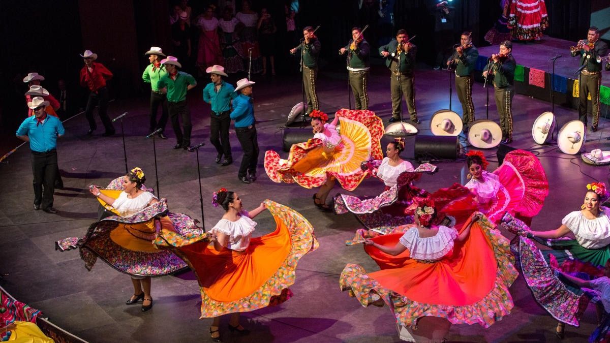 Mexican folk dancers in vibrant dresses perform on stage, musicians in background.